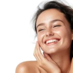 Woman smiling with hands on her face, showing radiant skin, surrounded by skincare products like cleansers and treatments
