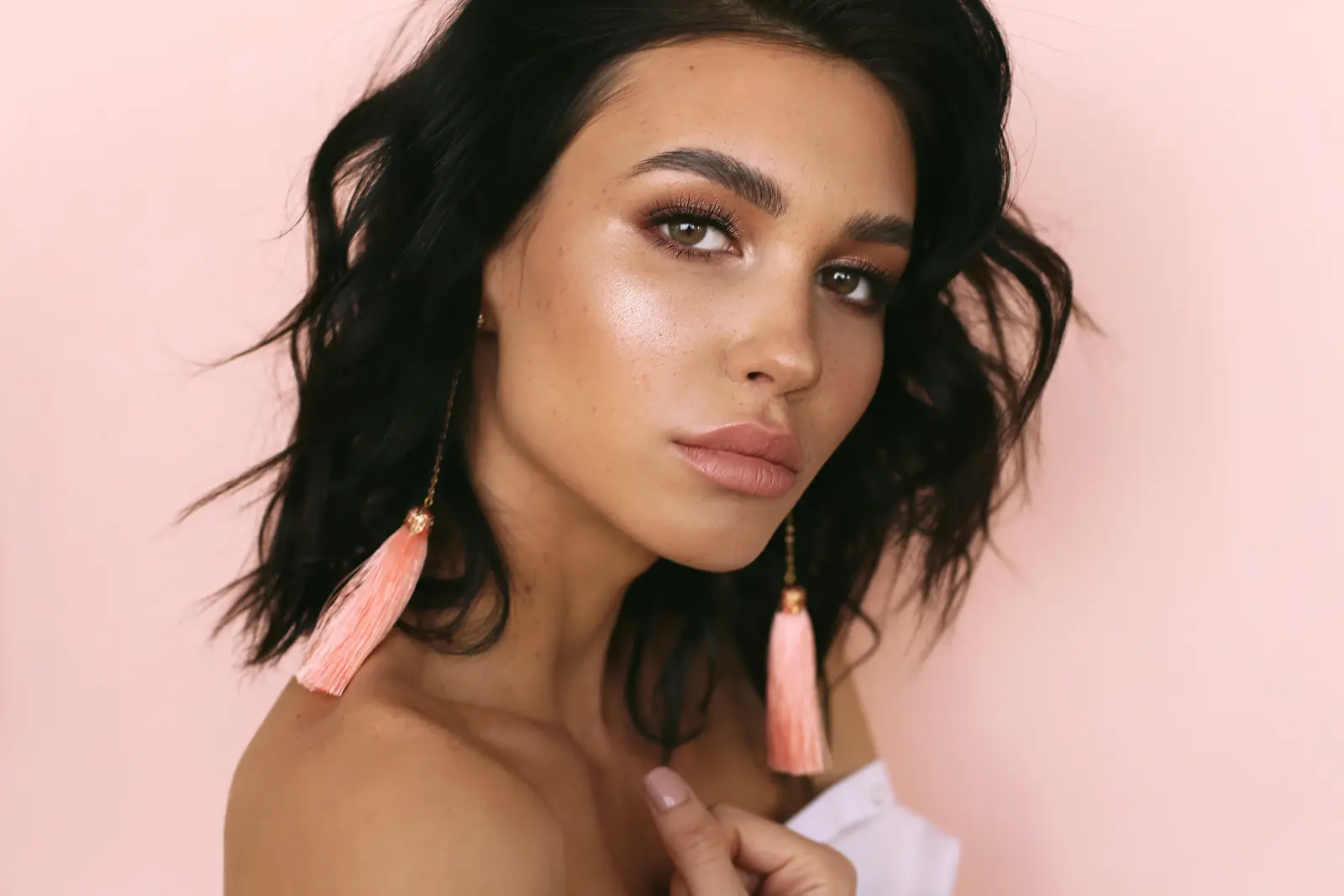 Woman with glowing skin and pink tassel earrings against a peach background.